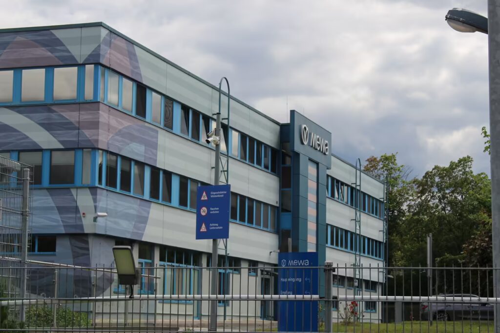 Bürogebäude der MEWA in Jena mit einem unverwechselbaren Design und einem eingezäunten Eingang; Ein berufliches oder unternehmerisches Umfeld; Das Gebäude verfügt über eine grau-blaue Fassade mit dem "Mewa"-Logo
