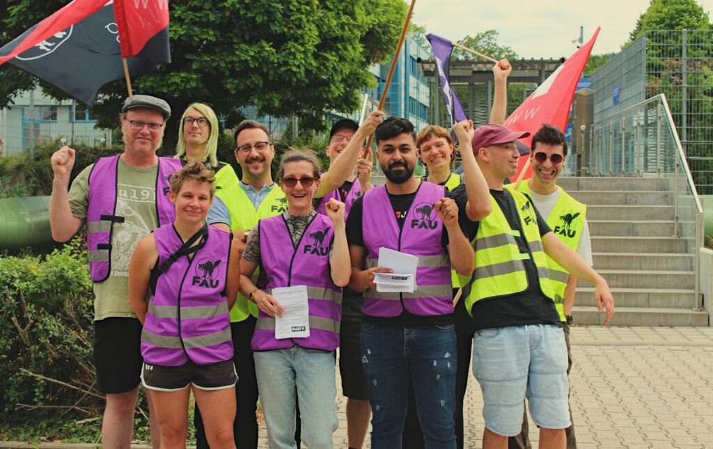 Eine Gruppe von Menschen, die meist lila oder gelbe Westen mit dem Aufdruck "FAU" tragen, posiert für ein Foto, während sie Fahnen und Fäuste in die Höhe strecken