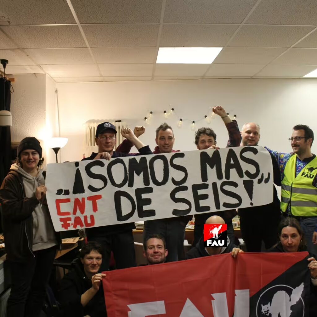 Zu sehen ist das innere eines Raumes. 6 Personen halten ein Banner hoch mit der Aufschrift "!SOMOS MAS DE SEIS!" in schwarzer Schrift. Links daneben steht in rot: "CNT FAU". Darunter sitzen 4 weitere Personen, die ein FAU Banner hochhalten.