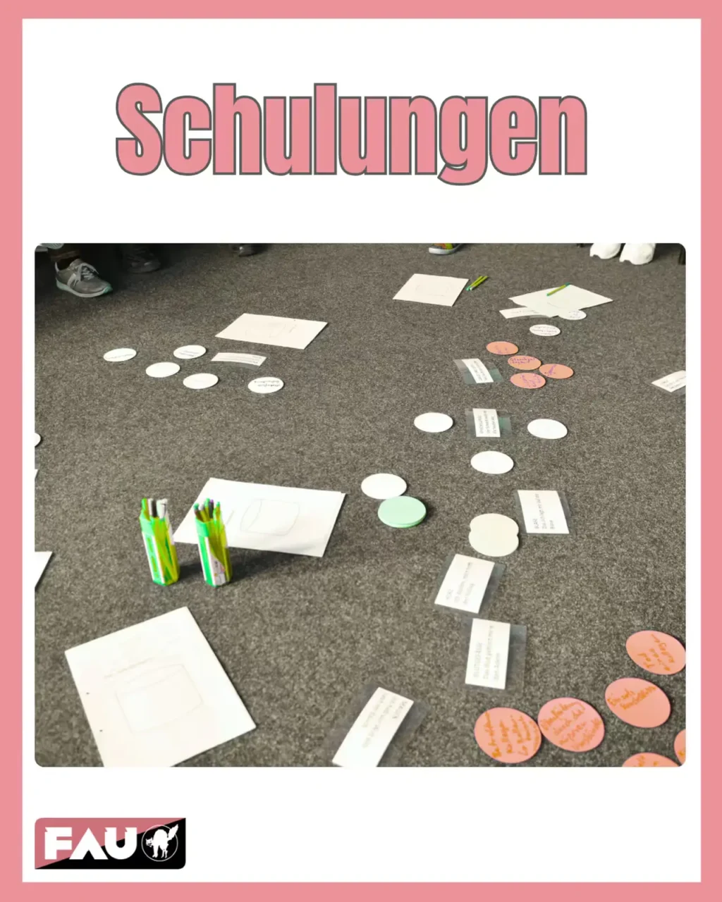 Grafik mit pinkem Außenrand und weißer Rahmenfläche: Oben steht in großer, rosa Schrift mit grauem Kontur der Begriff „Schulungen“. Die zentrale Fläche zeigt ein Foto: Auf einem dunkelgrauen Teppichboden liegen Materialien für eine Gruppenarbeit oder praktische Schulung verteilt: Mehrere weiße Blätter Papier mit leeren Umrissen, runde Sticker in Weiß, Rosa/Orange und Hellgrün (einige rosa Sticker sind handbeschriftet), zwei grün-gelbe Kassetten mit bunten Stiften sowie kleine, beschriftete Papierzettel in transparenten Hüllen. Am Rand des Fotos sind die Füße einiger Personen sichtbar – sie sitzen vermutlich in einer Gruppe auf dem Boden. Unten links ist das Logo der Friedrich-Alexander-Universität Erlangen-Nürnberg (FAU) sowie ein weiteres Logo mit einer Wildkatze abgebildet.
