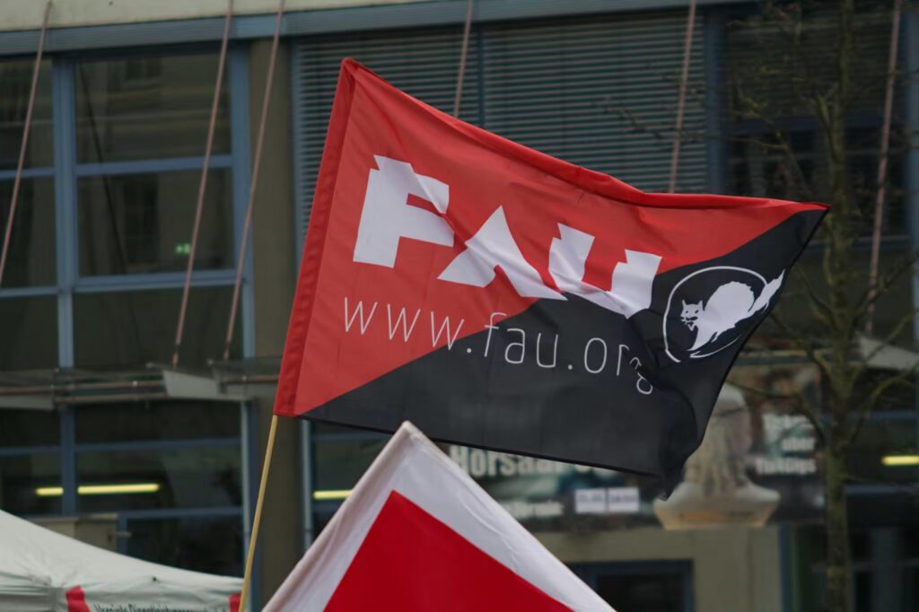 Eine rot-schwarze Flagge der Freien Arbeiterinnen- und Arbeiter-Union (FAU) weht im Wind vor dem Eingang zur Uni auf dem Abbe Platz in Jena. Die Flagge ist diagonal geteilt: Die obere linke Hälfte ist rot, die untere rechte Hälfte ist schwarz. Im Zentrum der Flagge stehen in großen, weißen Buchstaben die Initialen „FAU“. Darunter befindet sich in kleinerer, weißer Schrift die Internetadresse „www.fau.org“. Auf dem schwarzen Teil der Flagge, ganz rechts, ist das Logo der Gewerkschaft zu sehen: eine weiße, stilisierte, buckelnde Katze in einem weißen Kreis. Im unteren Bildrand ist ein Teil einer weiteren rot-weißen Flagge sowie ein Stück eines weißen Zeltes erkennbar. Der Hintergrund ist leicht unscharf und zeigt ein mehrstöckiges Gebäude mit großen Fensterfronten und blauen Metallstreben. Rechts im Bild ragt ein kahler Baum empor. Die gesamte Szene wirkt wie eine Aufnahme von einer Kundgebung oder Demonstration im Freien.
