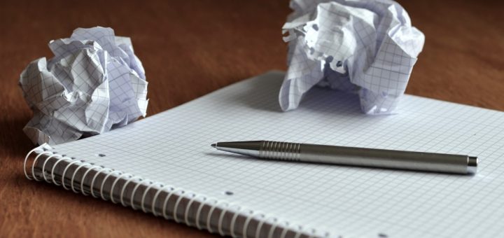 Nahaufnahme: Spiralblock mit kariertem Papier auf dunklem Holzschreibtisch. Auf dem Block liegt ein silberner Kugelschreiber. Zwei fest zerknüllte Blätter karierten Papiers stehen daneben.